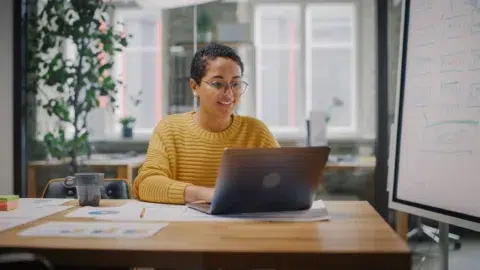 A person wearing glasses and a yellow sweater smiles while working on a laptop at a community bank Massachusetts office filled with papers in a bright, modern space.