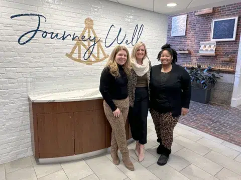 Three women smiling and standing together indoors in front of a white brick wall that says Journey Well—celebrating success in bank jobs.