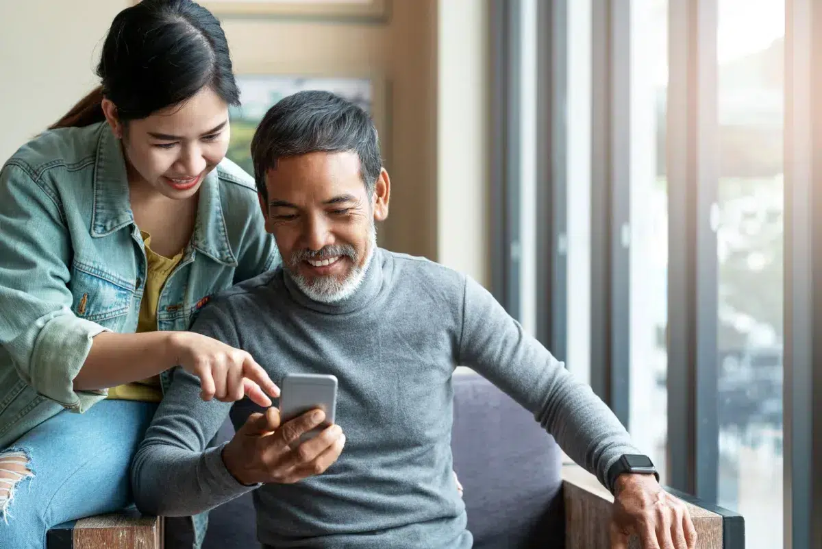 A smiling woman leans over a seated man as they both explore secure mobile banking on a smartphone together by a large window in Massachusetts.