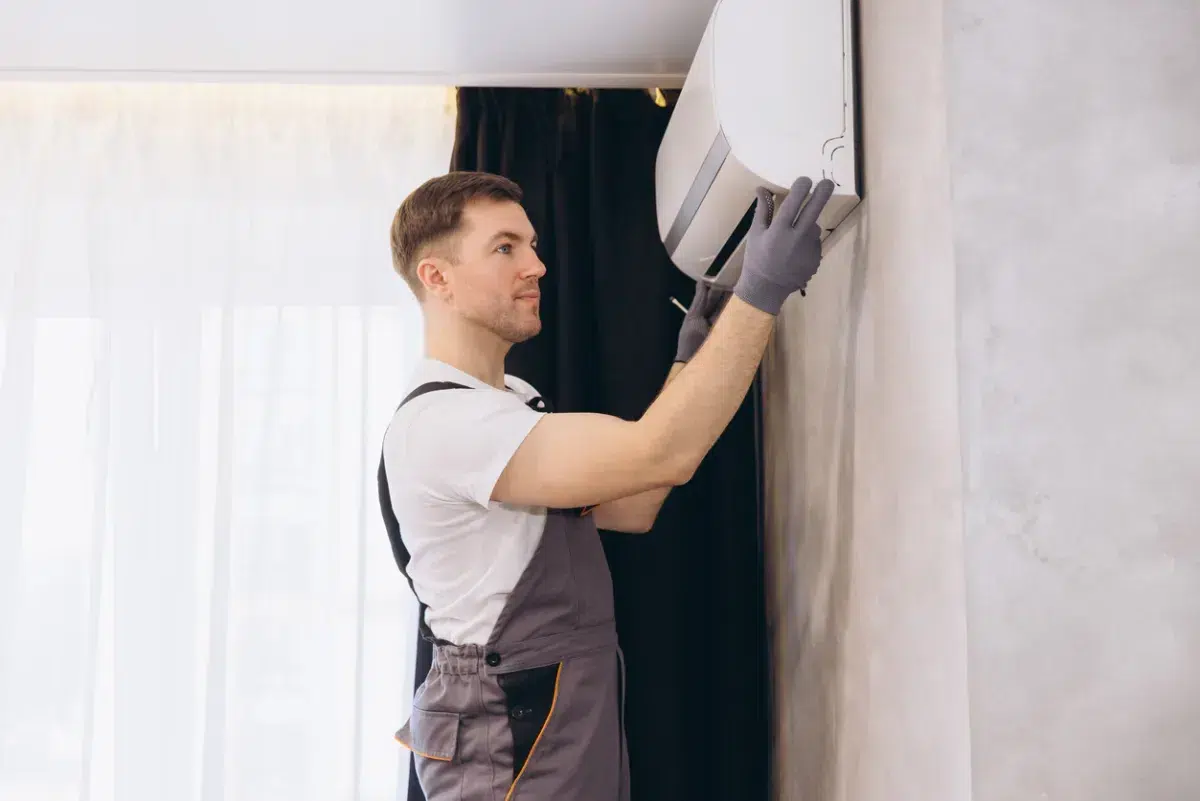 A technician in gray gloves and overalls services a wall-mounted air conditioner, which may qualify for a Mass Save heat loan, in a room with light curtains.