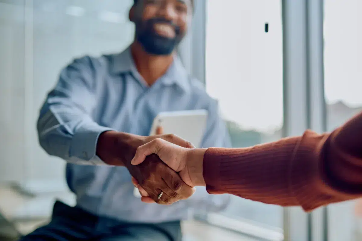A man in a blue shirt smiles while shaking hands with another person in a brown sweater at a community bank in Massachusetts, sitting indoors by a window.