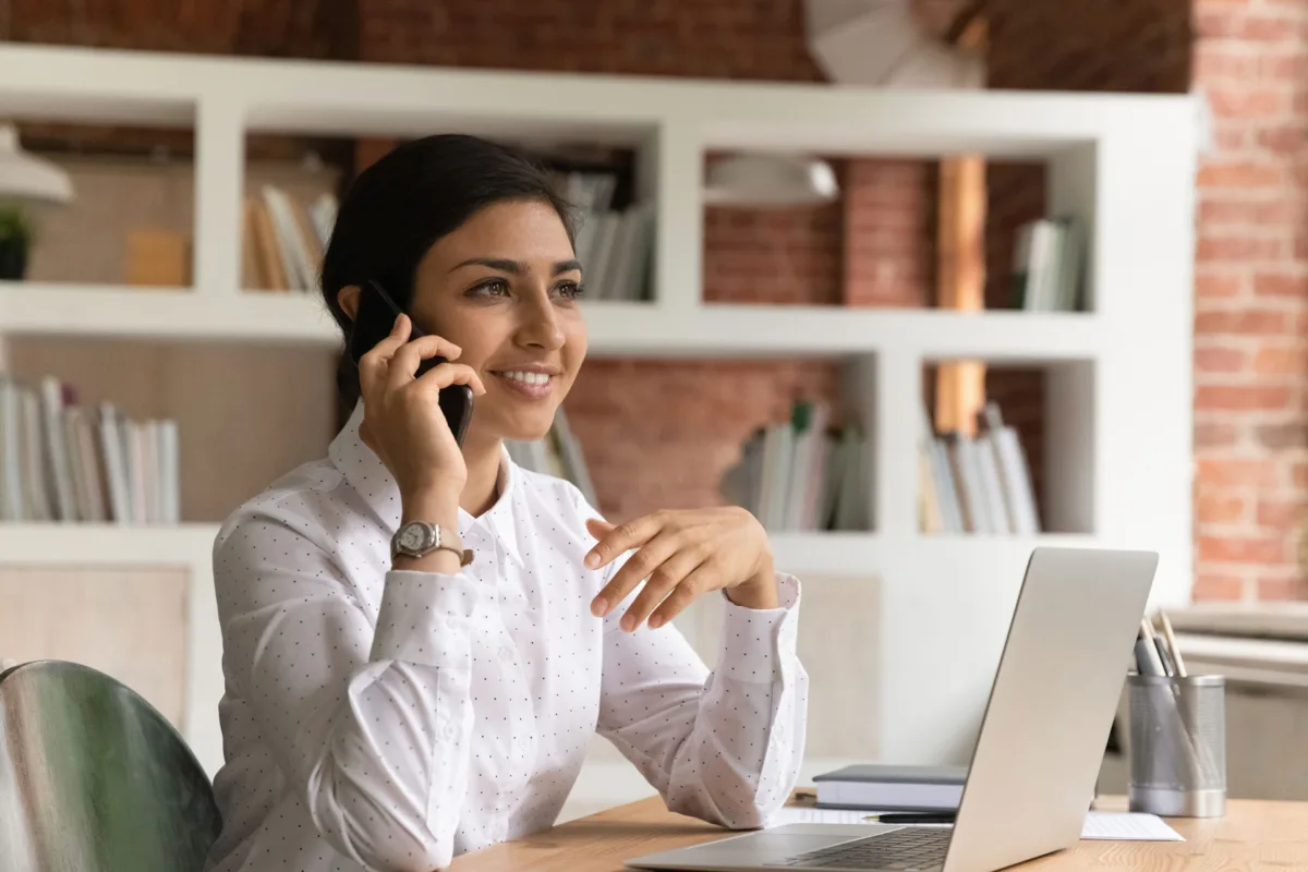 A woman sits at her desk in a modern office, smiling while using secure mobile banking Massachusetts on her smartphone, with an open laptop and shelves of books behind her.