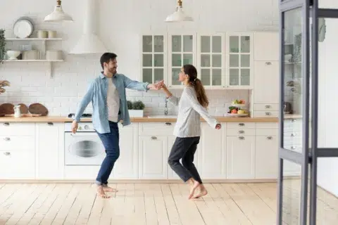 A man and woman dance barefoot together in their bright, modern kitchen—perhaps dreaming of a home made possible by a 40 year mortgage. White cabinets and fruit complete the scene.