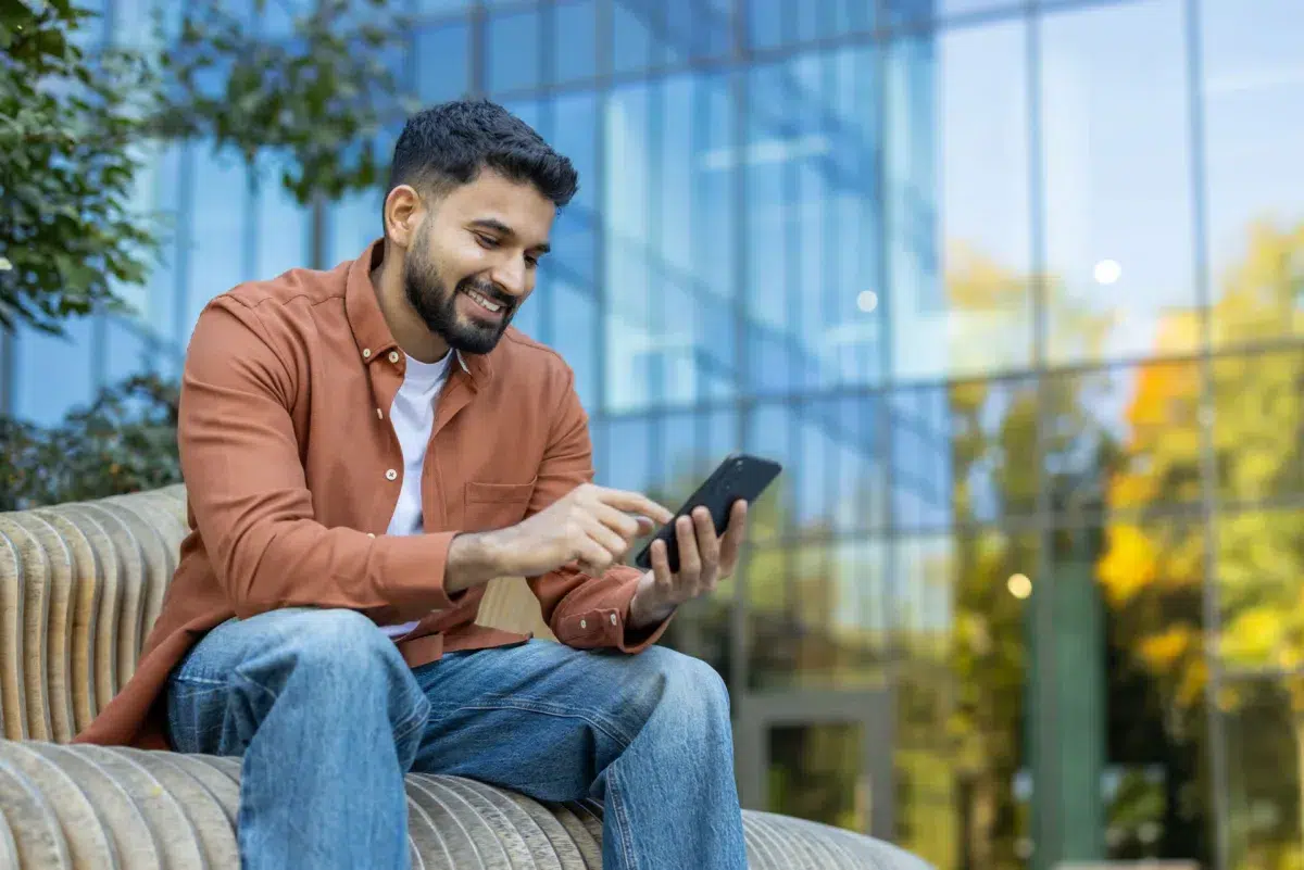 A man sitting outdoors on a curved bench in Massachusetts, smiling and using secure mobile banking on his smartphone, with a modern glass building in the background.