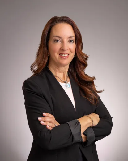 A woman with long brown hair in a black blazer poses with arms crossed, smiling against a gray background.