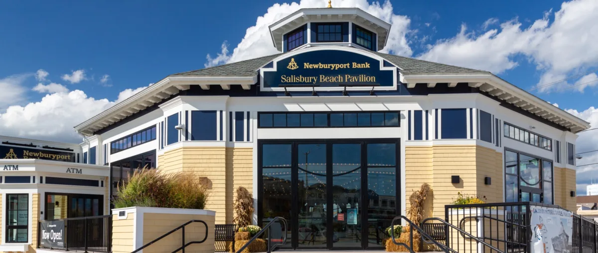 Octagonal building with large glass doors, beige and blue exterior, and signs reading Newburyport Bank and Salisbury Beach Pavilion under a bright blue sky.