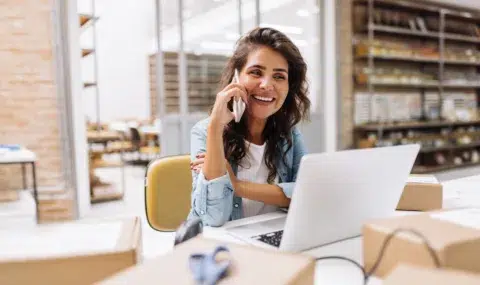 A smiling woman sits at a desk with a laptop, talking on the phone, surrounded by cardboard boxes in a modern workspace.