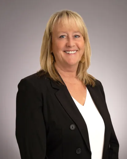 A woman with blonde hair wearing a black blazer and white blouse smiles at the camera against a plain gray background.