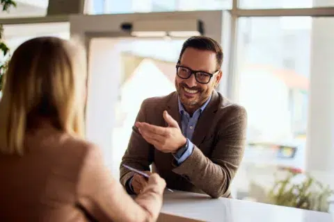 Man in glasses and a brown blazer smiling and discussing safe deposit boxes with a woman across a desk in a bright office setting.