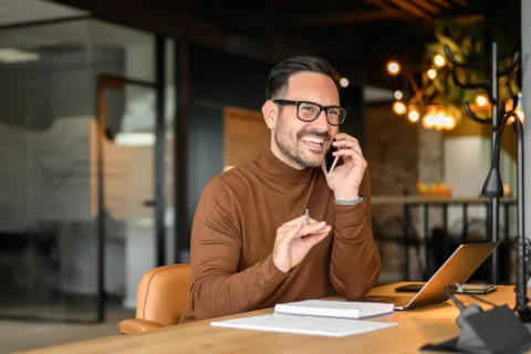 Smiling man in a brown turtleneck talks on the phone at a desk with a laptop, notebook, and pen in a modern office setting.