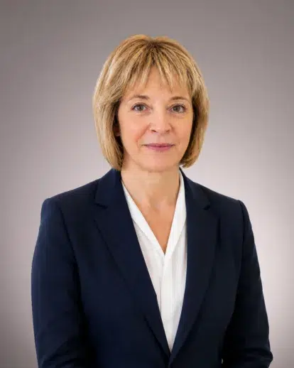 Jodi Panzica, wearing a dark blazer and white blouse, stands against a plain gray background, facing the camera and smiling slightly.
