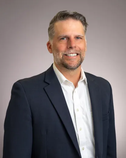Scott Beattie wearing a dark suit jacket and white shirt, standing in front of a plain light gray background.