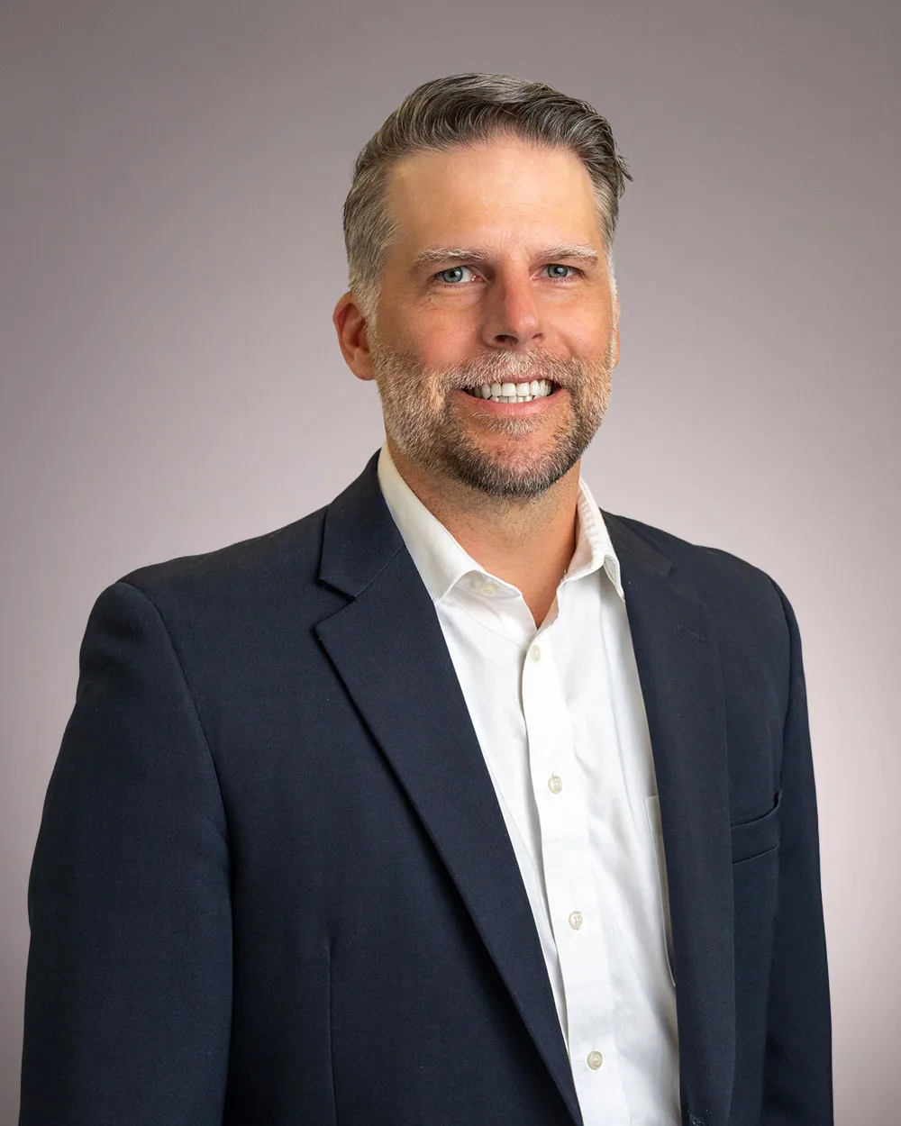 Smiling man with short gray hair and beard wearing a dark suit jacket and white shirt, standing in front of a plain light gray background.