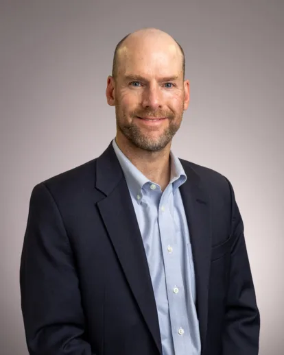 A man in a dark suit and light blue shirt smiles at the camera, standing against a plain light gray background.