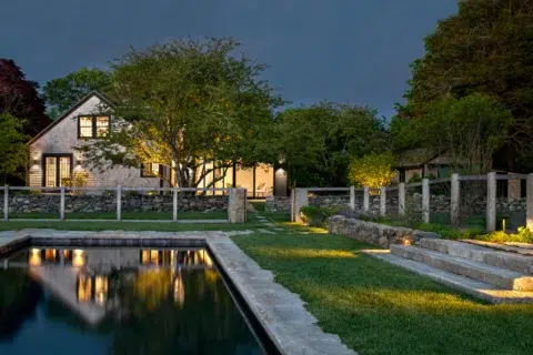 A stone house with large windows and exterior lights, surrounded by trees and a stone fence, sits beside a rectangular pool at dusk.