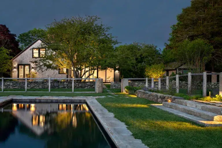 A stone house with large windows and exterior lights, surrounded by trees and a stone fence, sits beside a rectangular pool at dusk.