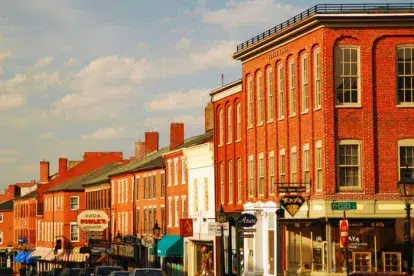 A row of historic brick buildings with shops and restaurants on a sunny day, cars parked along the street, and a blue sky with clouds above.