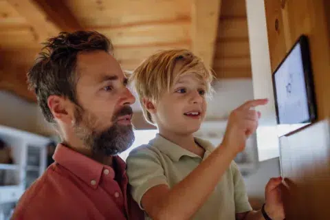 A man holds a young boy who is touching a digital screen, exploring home upgrades with Mass Save Heat Loan info, inside a cozy wooden room.