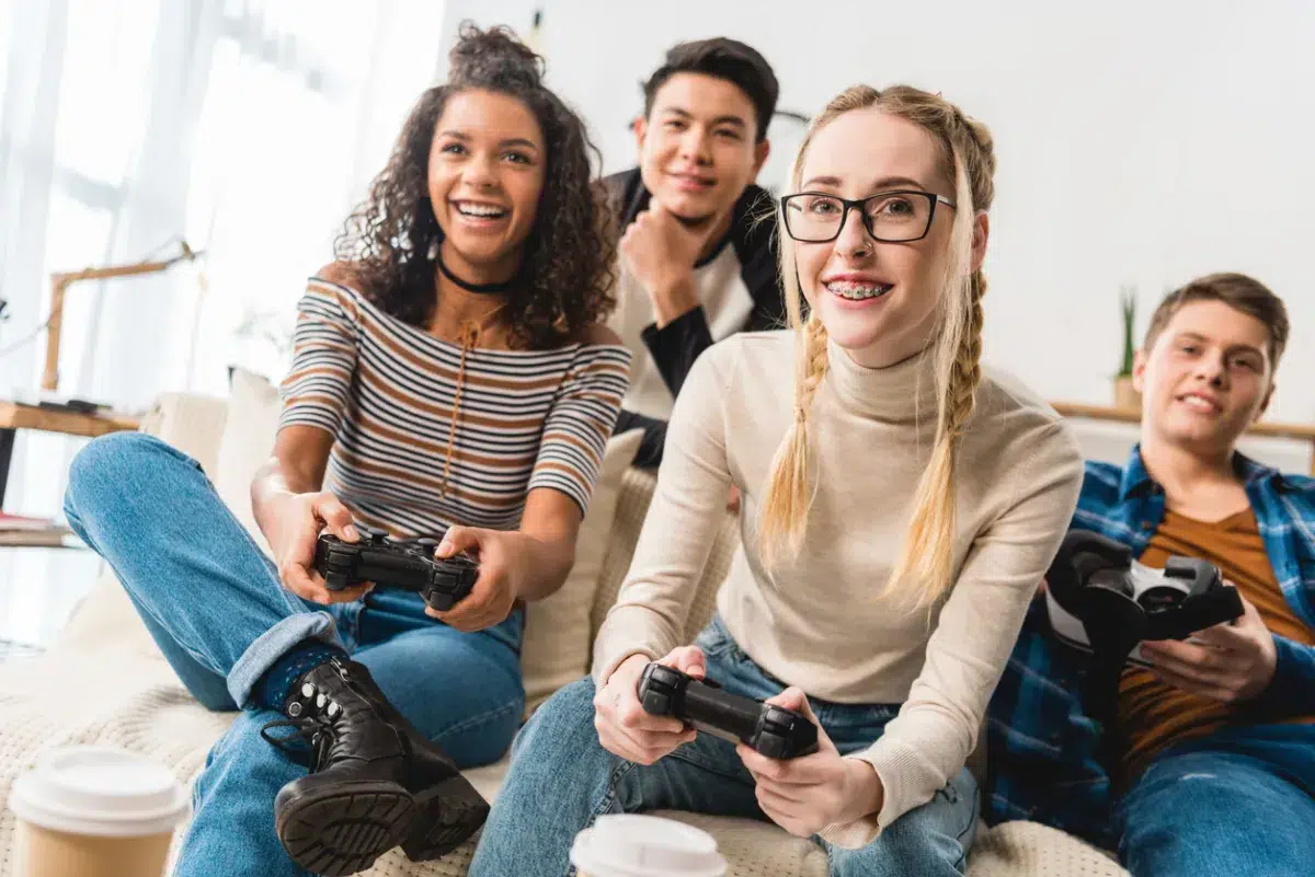 Four smiling teenagers sit on a couch playing video games together, holding controllers, with coffee cups and a Mass Save Heat Loan brochure on the table in front of them.