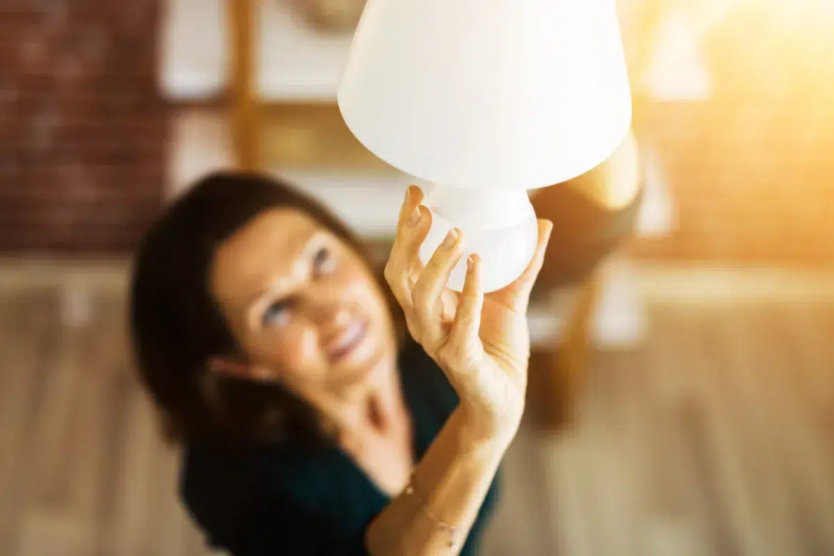 A woman reaches up to change a lightbulb in a ceiling lamp, smiling as sunlight streams in, inspired by savings from a Mass Save heat loan.