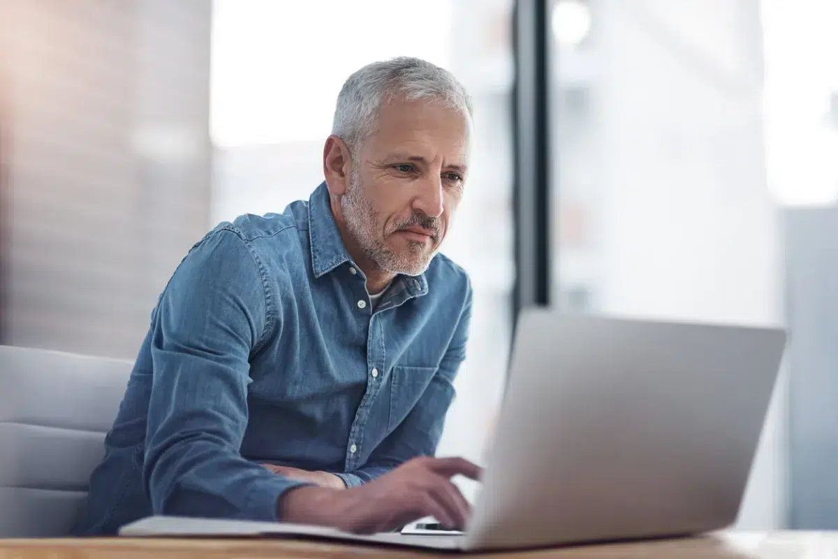 Older man with gray hair and beard, wearing a denim shirt, sits at a desk using his laptop to review banking resources in a bright, modern office setting.